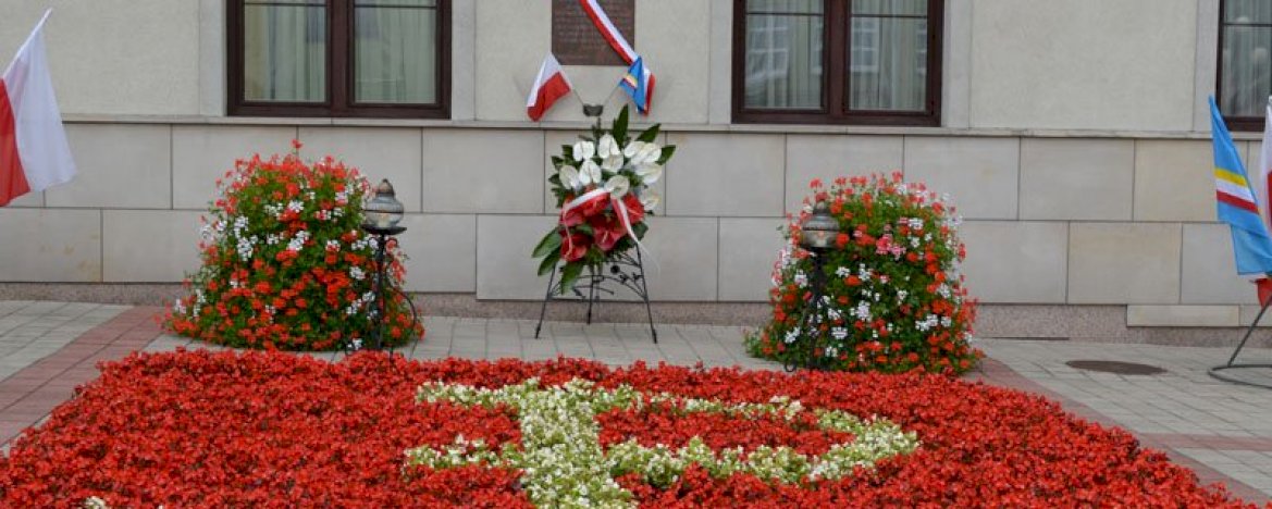 Tablica zawieszona na scianie budynku, z przewieszoną biało-czerwoną wstęgą, pod spodem wiązanka kwiatów, kwiaty na pierwszym planie, układające się w znak PW