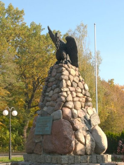 Zdjęcie pomnika , sttos spojonych kamieni, z przymocowaną, czarna tablicą, na nim metalowy orzeł trzymający w dziobie łańcuch, w tle drzewa