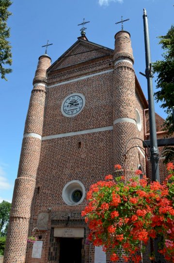 Kościów w Serocku,  ceglana fasada z dwiema kolumnowymi wieżami po bokach i dużym zegarem na środku, cegły z białymi wstawkami. Na dole kwiaty w donicy,obok latarnia. Nad zegarem znajdują się krzyże, a niebo w tle jest błękitne.