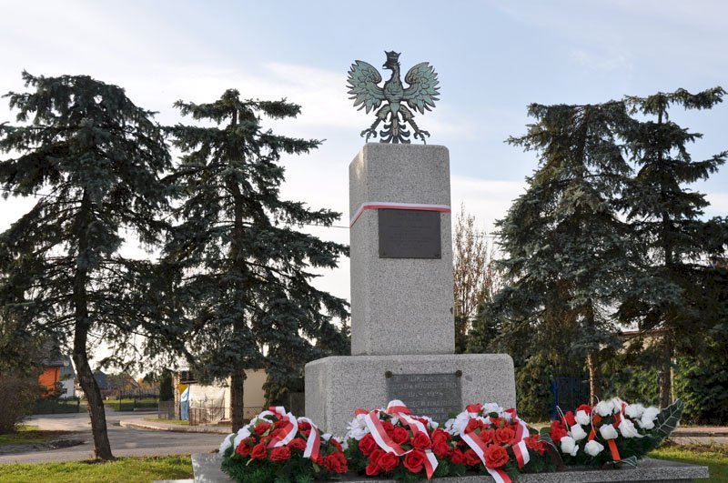 Pomnik z dwóch olbrzymich bloków kamiennych, metalowy orzeł na szczycie, na nim 2 tablice, pod nim kwiaty, za nim drzewa